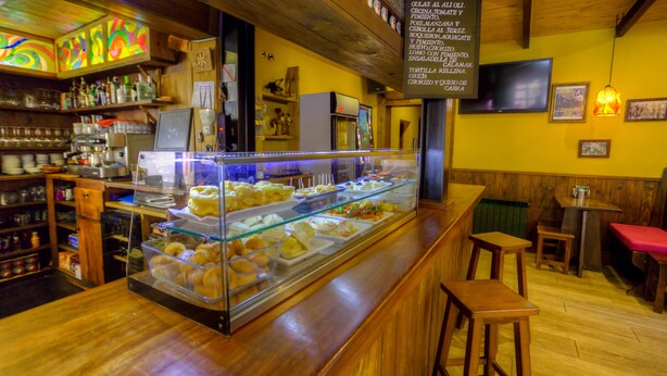 A bakery counter with a variety of baked goods, a coffee machine, and a menu board listing items. - La Tintoreria (Villablino)