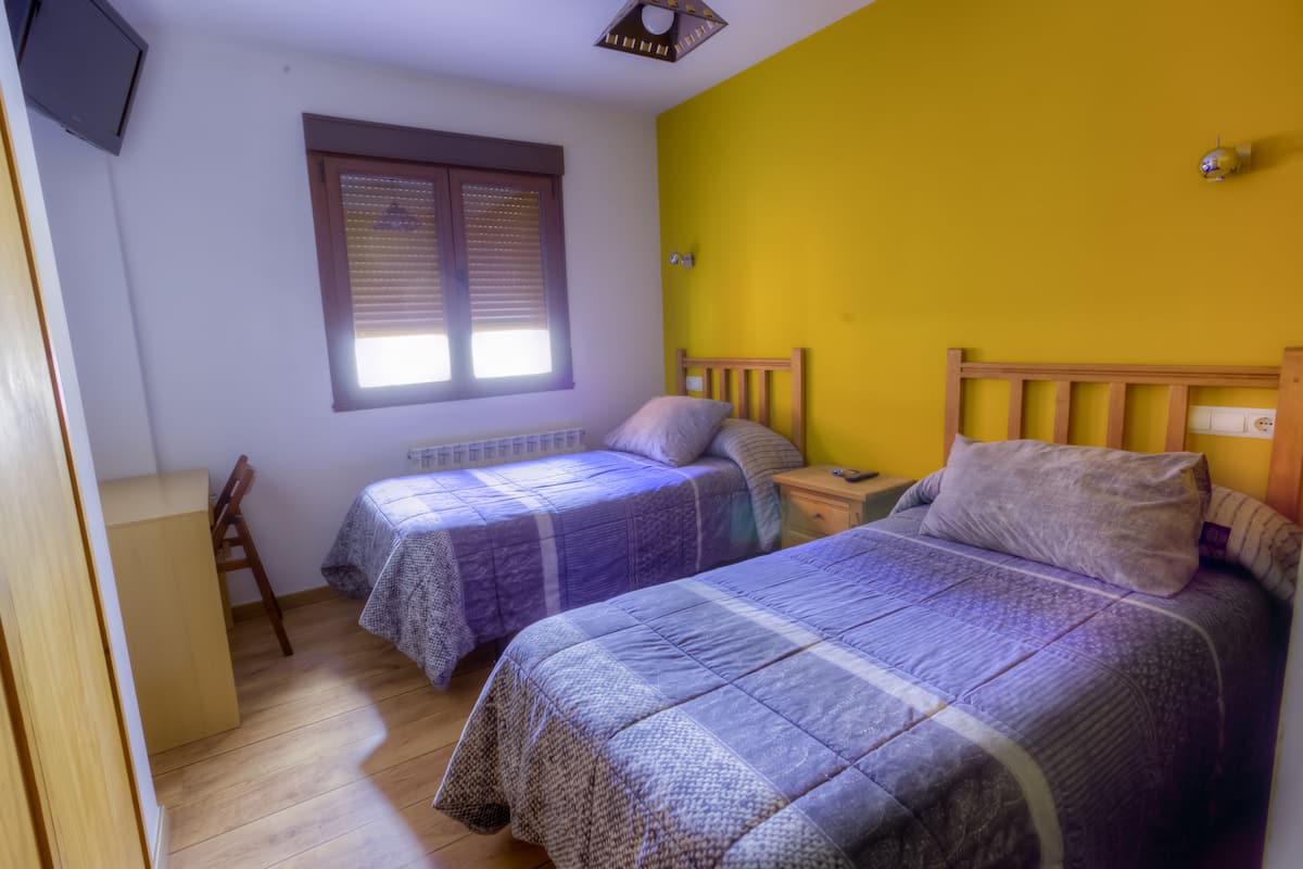 A bedroom with two beds, a window with blinds, a desk, and a small table.