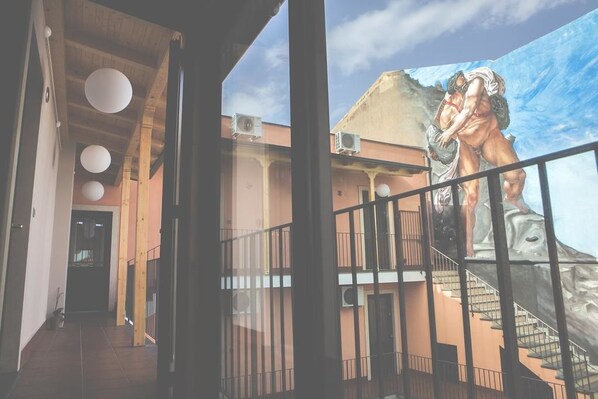 A corridor with a mural of a muscular figure on the wall, a staircase with metal railings, and a ceiling with wooden beams and spherical lights.