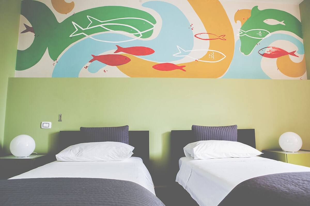 A hotel room with two beds, a colorful abstract mural on the wall, and a green wall.