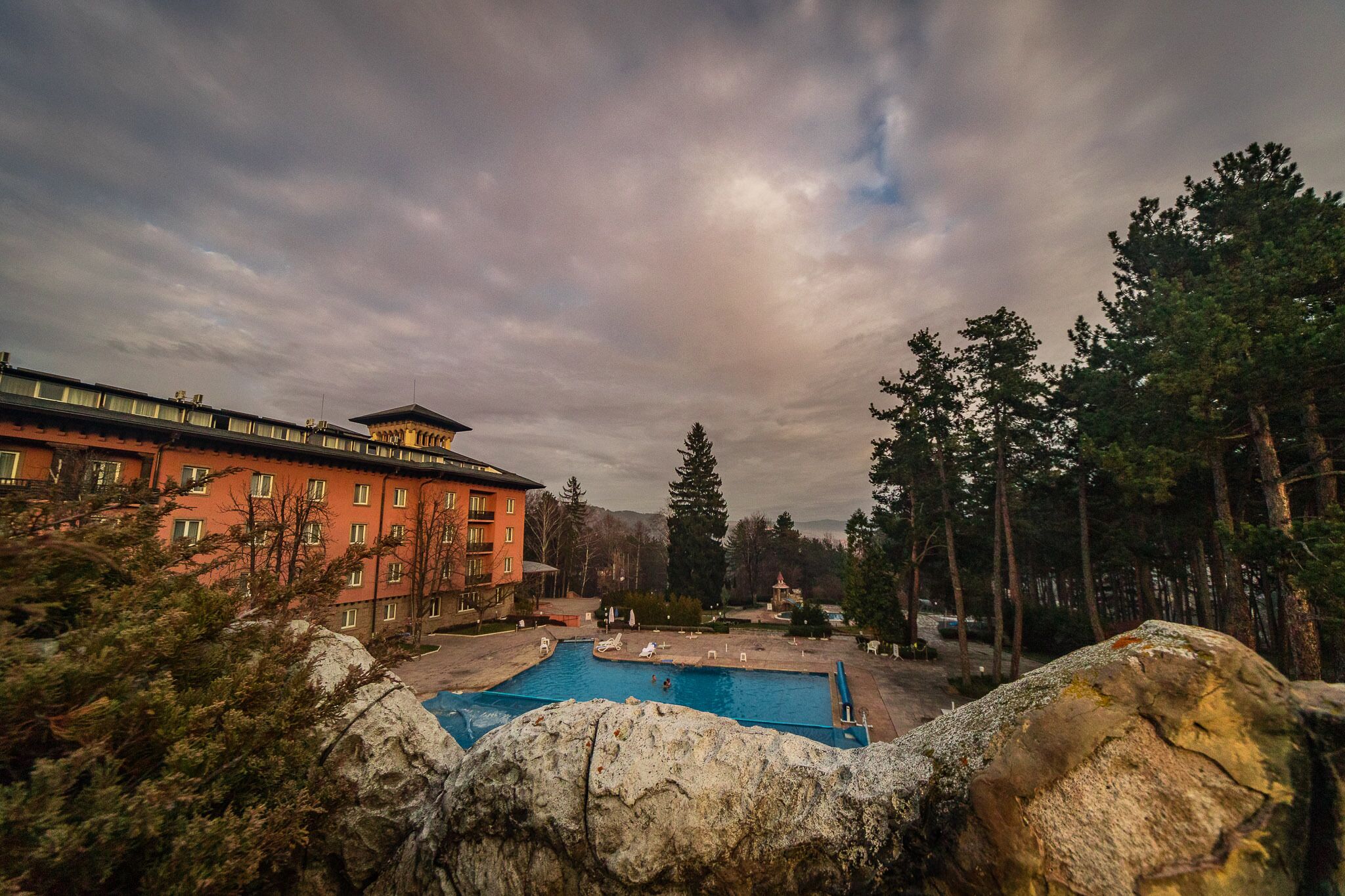 A hotel with a swimming pool surrounded by trees and a large rock in the foreground.