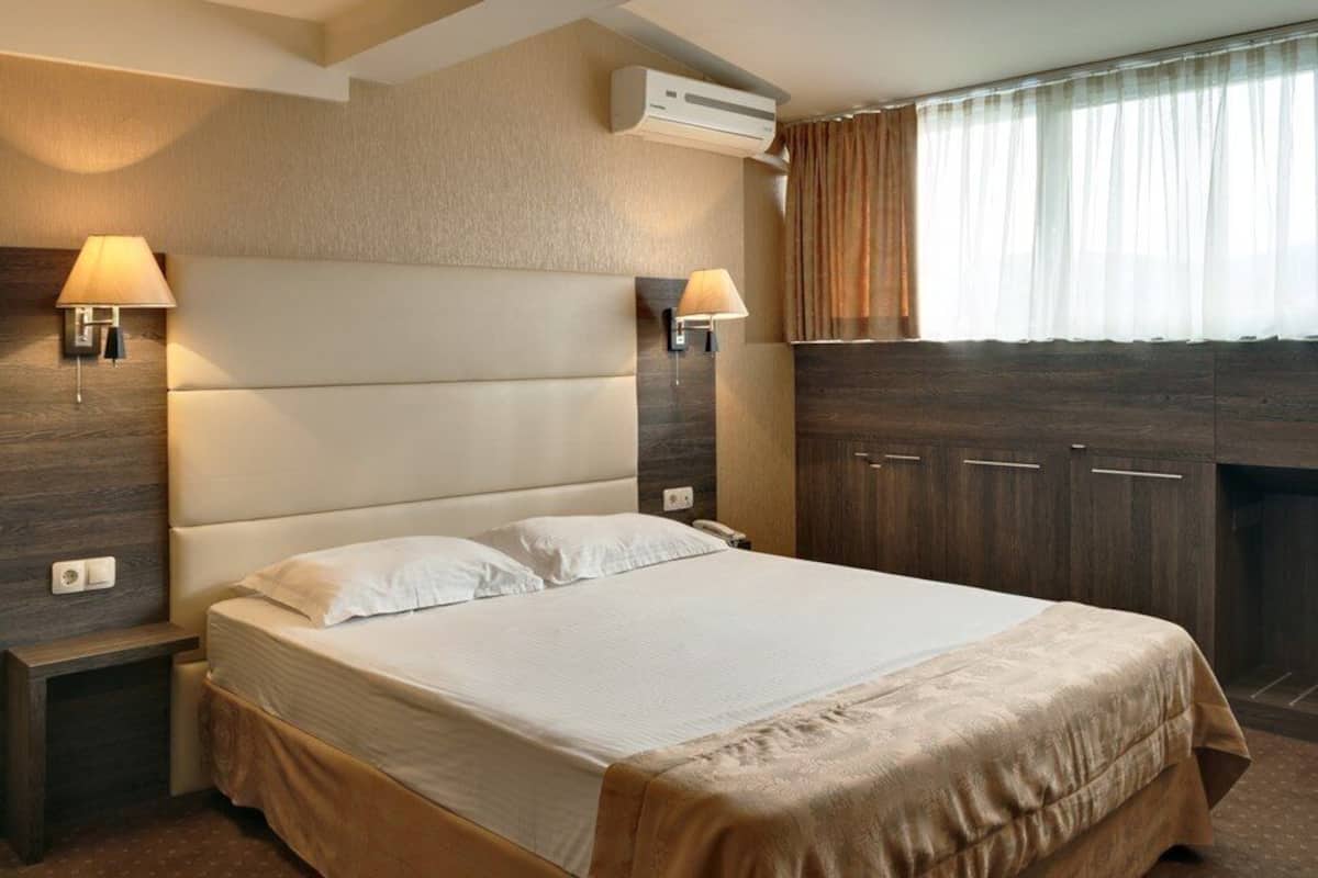 A hotel room with a large bed, two wall-mounted lamps, a wardrobe, and an air conditioning unit.