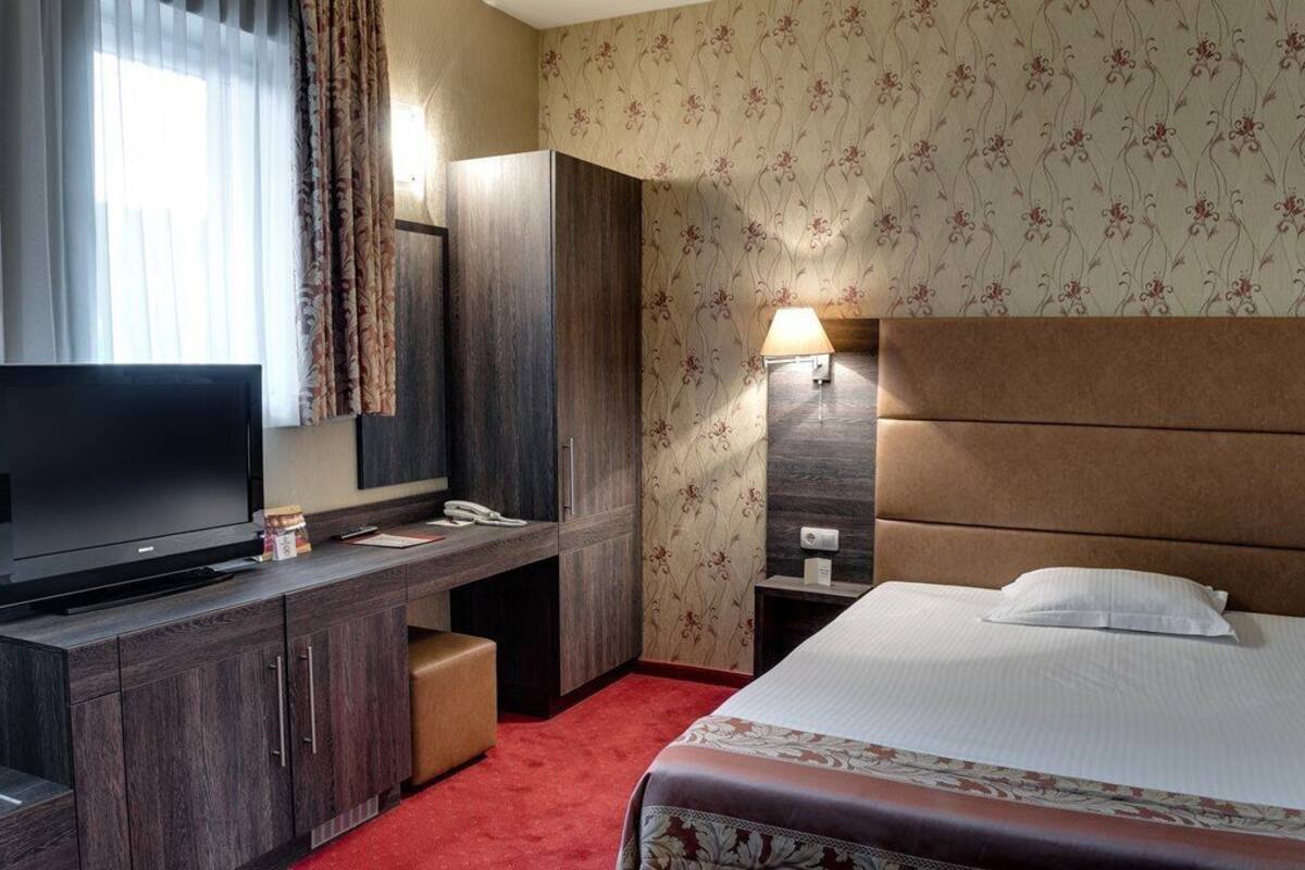 A hotel room with a large flat-screen TV, a wooden desk, a comfortable bed, and a nightstand with a lamp.