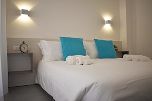 Un lit bien fait, avec des oreillers blancs et bleus, une horloge sur une table de chevet et deux lampes fixées au mur.