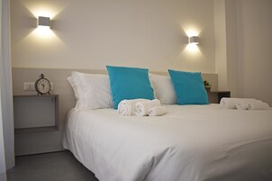 Un letto ben rifatto con cuscini bianchi e blu, una sveglia su un comodino e due luci a parete.