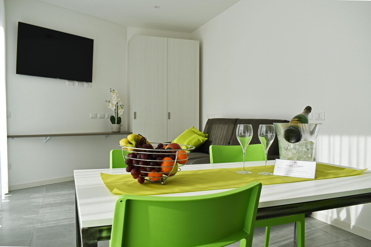 Un coin repas avec une table blanche, des chaises vertes, un panier de fruits et un verre à vin.