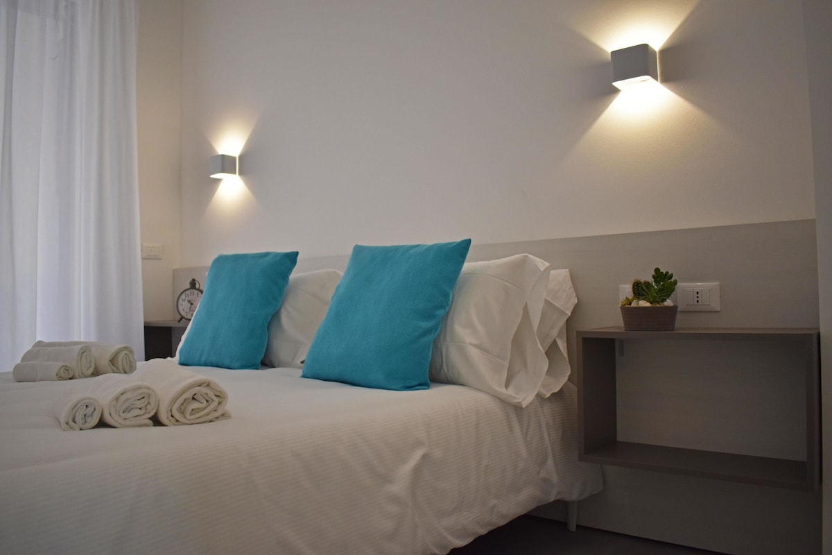 Un lit bien fait, avec des oreillers blancs et bleus, une table de chevet ornée d’une plante et un luminaire fixé au mur.