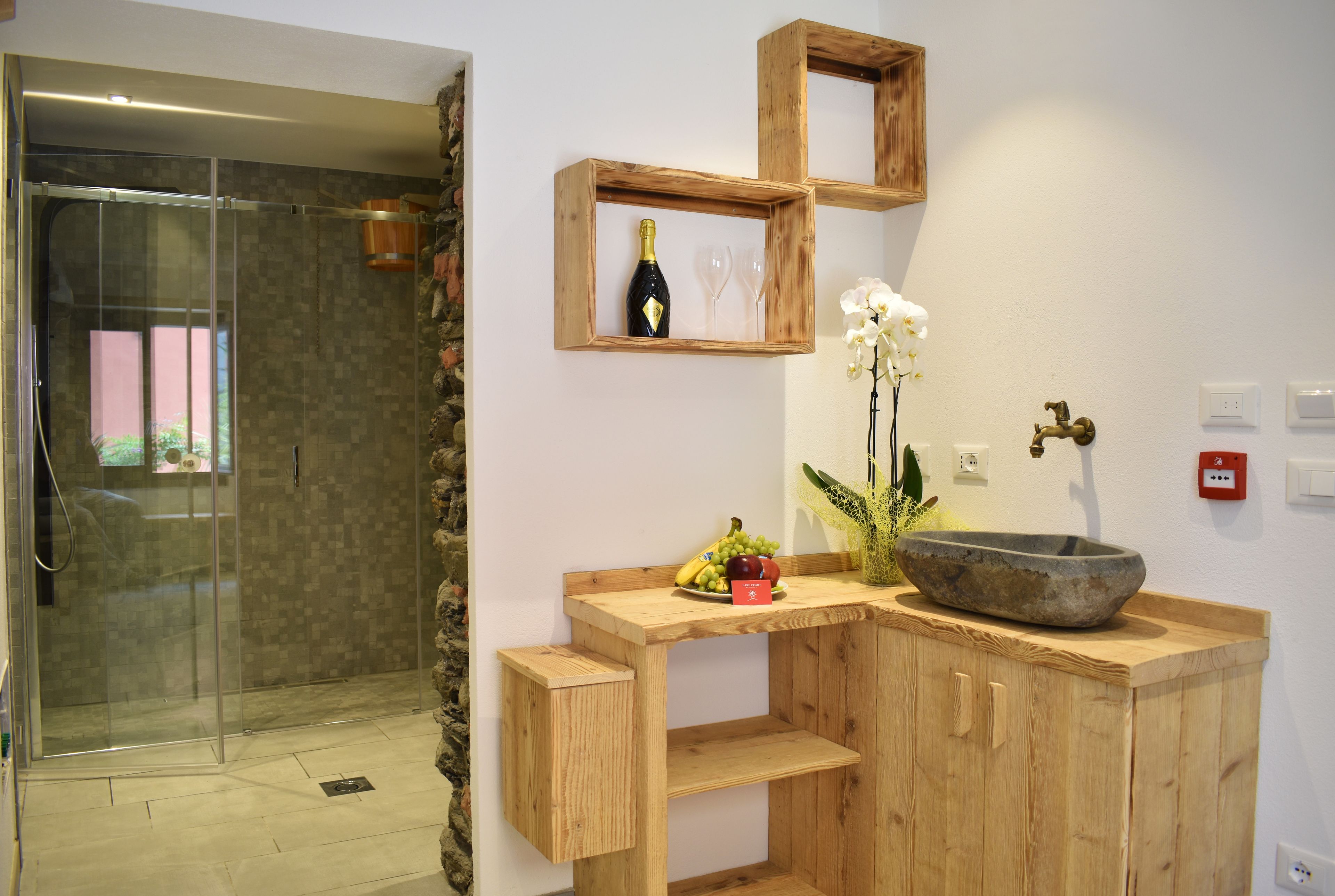Ein Badezimmer mit einer Duschkabine aus Glas, Holzregalen, einem Waschbecken mit Steintonne und einer Vase mit Blumen.