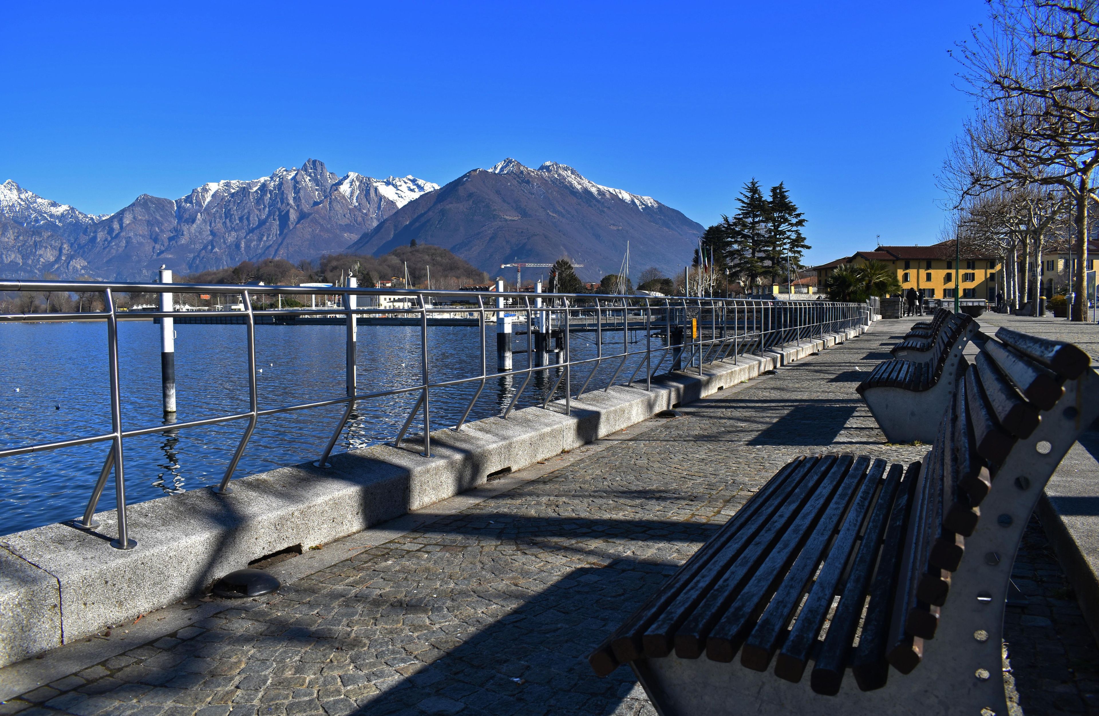 A waterfront promenade with benches, a metal railing, and a mountain backdrop.