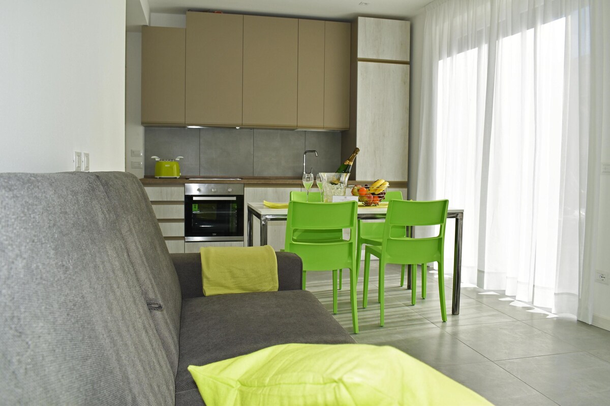 Un salon moderne avec un canapé gris, des fauteuils verts et une cuisine en arrière-plan.