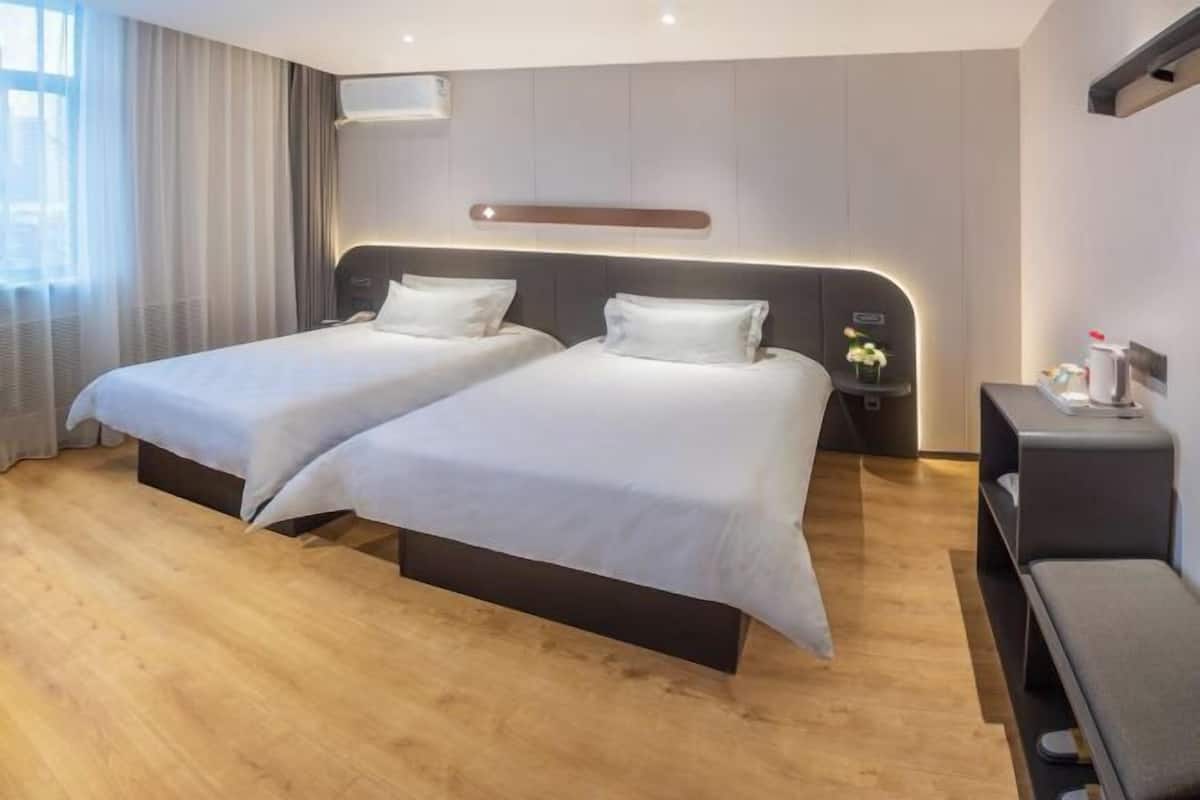 Habitación de hotel con dos camas, una mesita de noche con lámpara, una mesita auxiliar pequeña y una ventana con cortinas.