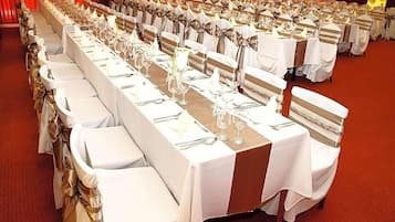 Un salón de banquetes con largas mesas dispuestas para un evento formal, cada una con manteles blancos y fajines de plata.