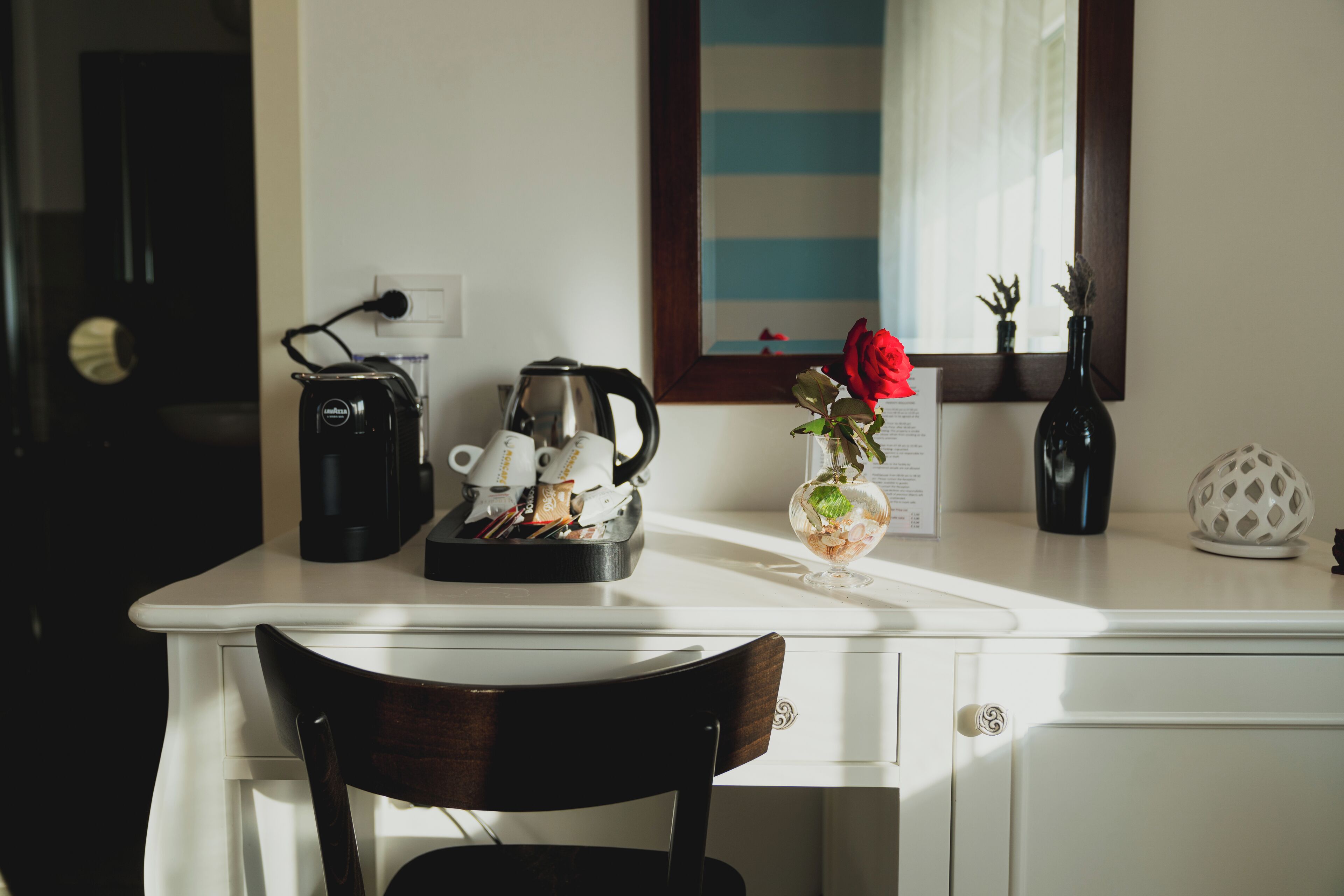 A white desk with a coffee maker, a kettle, and a vase with a red rose.