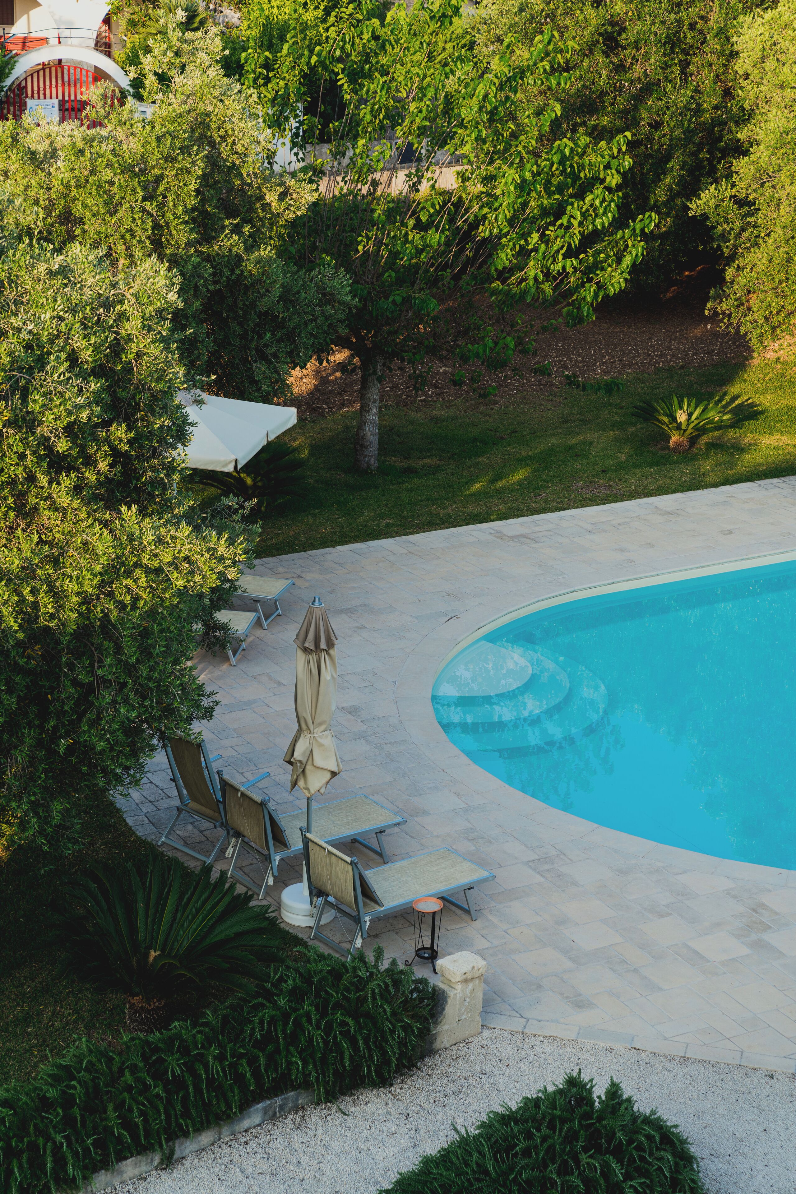 Piscina com borda curva, rodeada por um deck pavimentado. Há cadeiras de espreguiçadeira e um guarda-sol coberto.