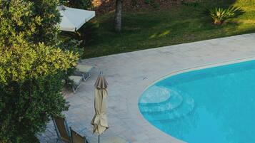 Piscina com borda curva, rodeada por um deck pavimentado. Há cadeiras de espreguiçadeira e um guarda-sol coberto.