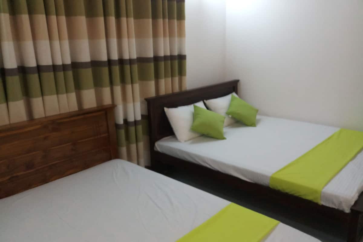 A room with two single beds, each with a wooden headboard and white bedding. There are two green pillows on one bed. The room has a window with curtains, and the walls are plain white.