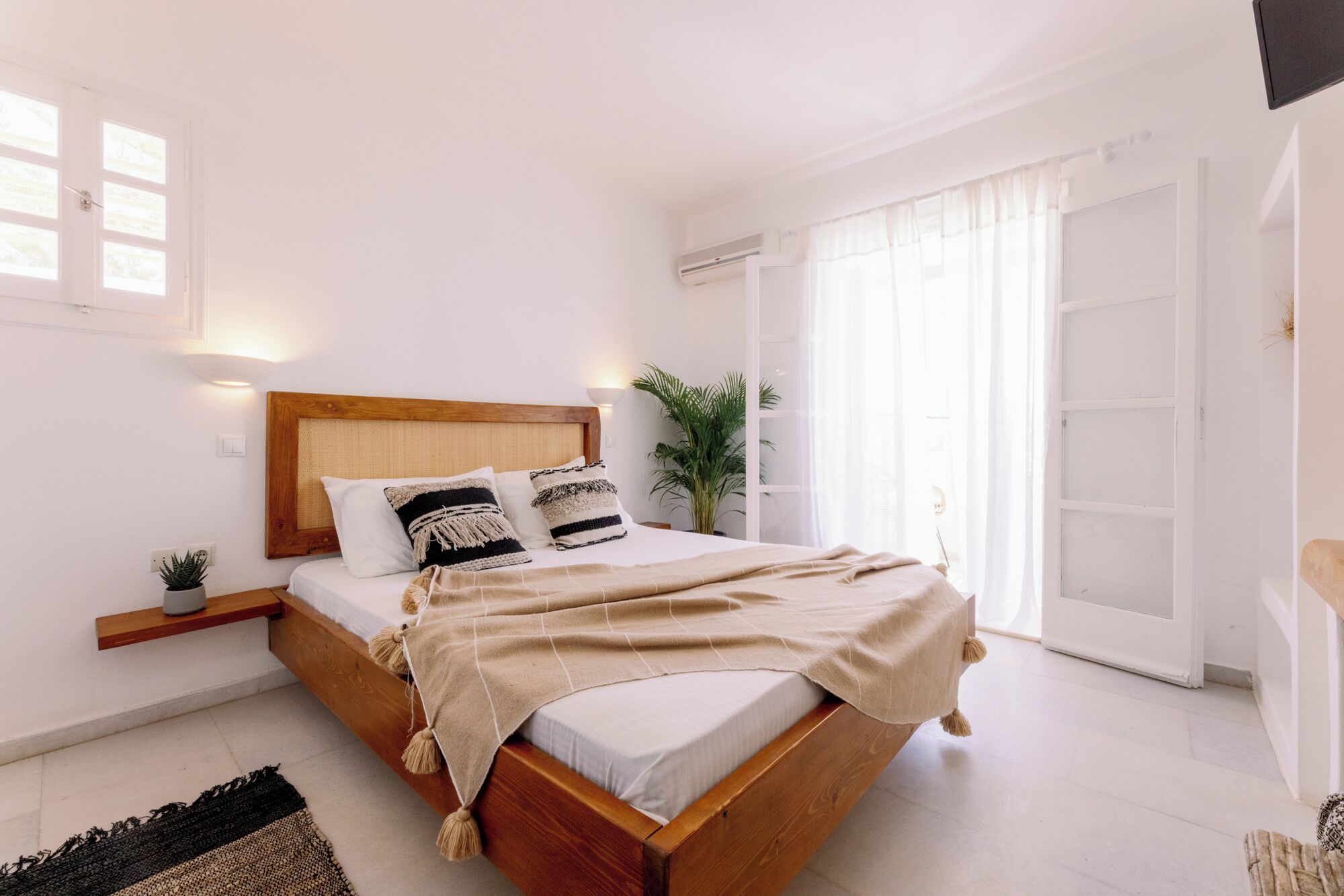 A bedroom with a wooden bed, white walls, a small plant, and a large window with curtains.