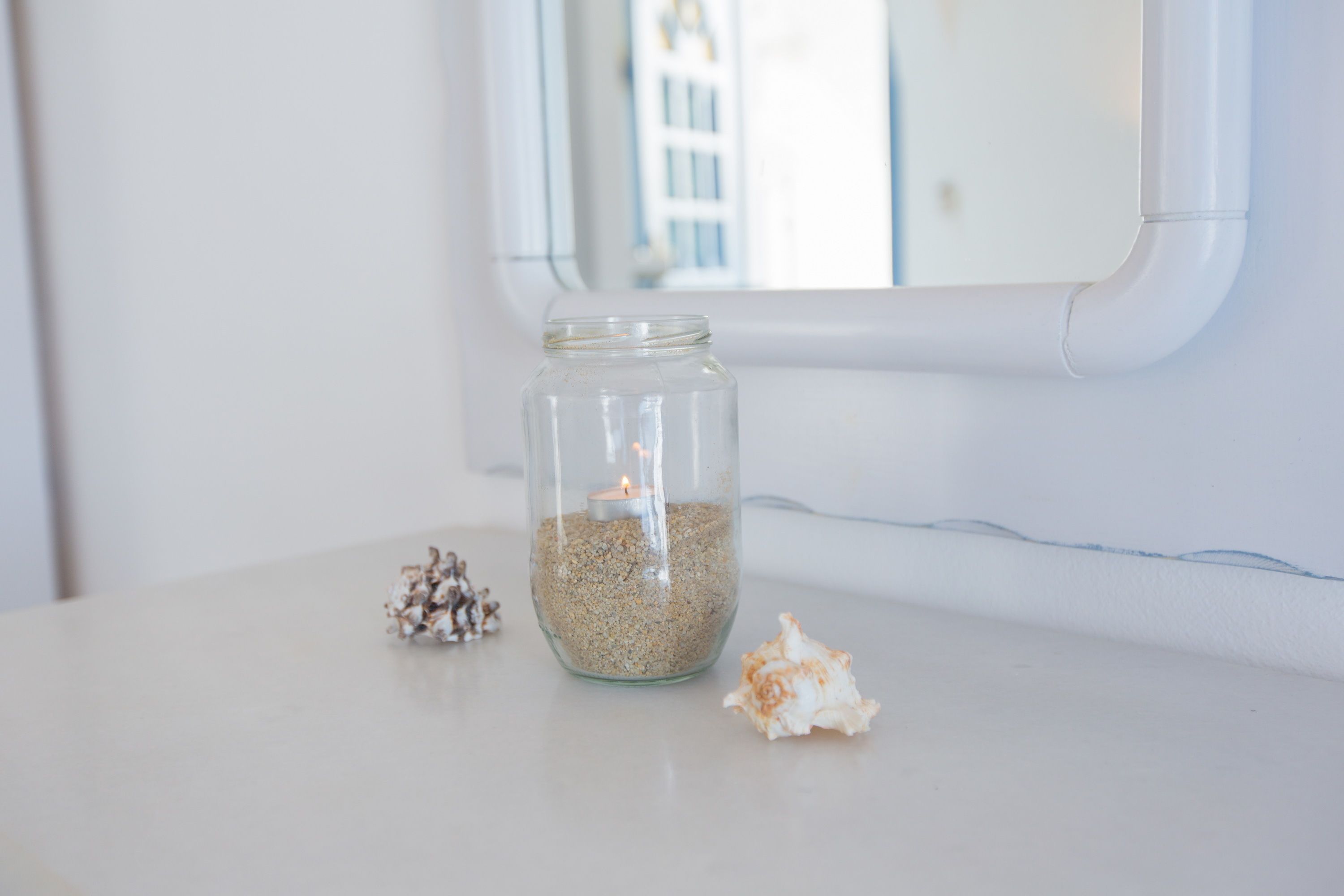 A glass jar filled with sand and a candle, placed on a white surface with a small seashell beside it.