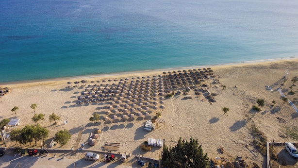 Vista aèria d'una platja amb files de papereres i gandules, una zona d'aparcament i una vista clara del mar i muntanyes llunyanes.