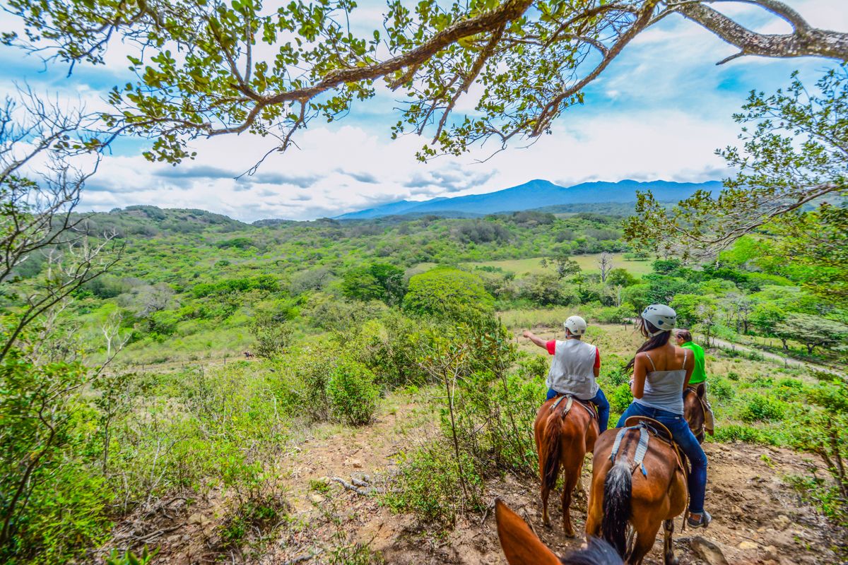 Two people riding horses on a dirt path with a view of a green landscape and mountains in the distance.