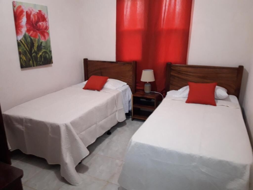 A hotel room with two single beds, each with a white bedspread and a red pillow. There is a nightstand with a lamp between the beds.