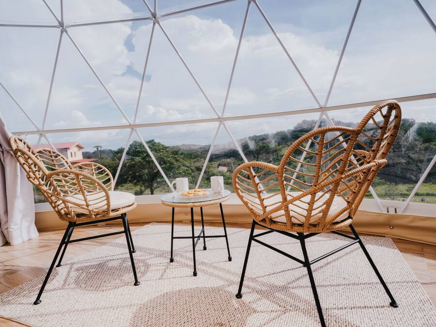 Quarto com uma grande cúpula de vidro, cadeiras de vime, uma mesa pequena e vista para uma paisagem verde.