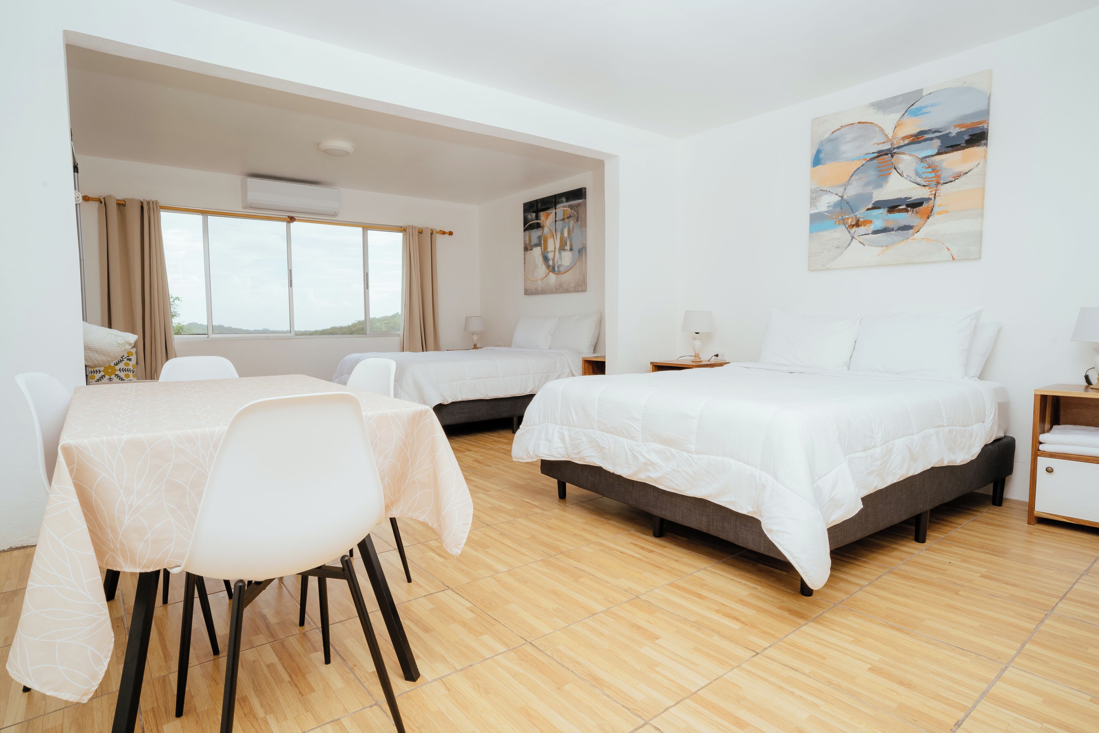 A hotel room with two beds, a dining table, chairs, a painting, and a view of the outdoors.
