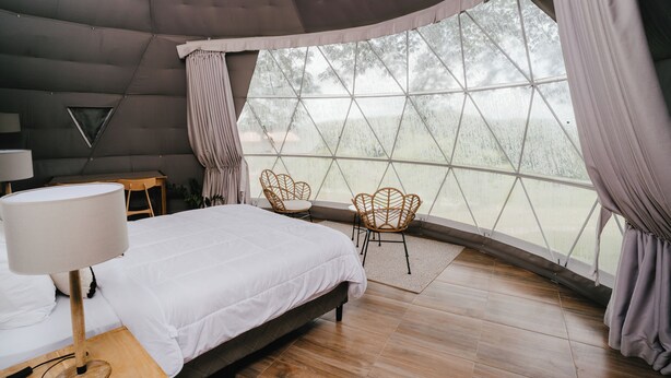 A bedroom with a bed, a nightstand, a lamp, a window with curtains, and two wicker chairs. - Vida Aventura Ranch (Mogote de Bagaces)