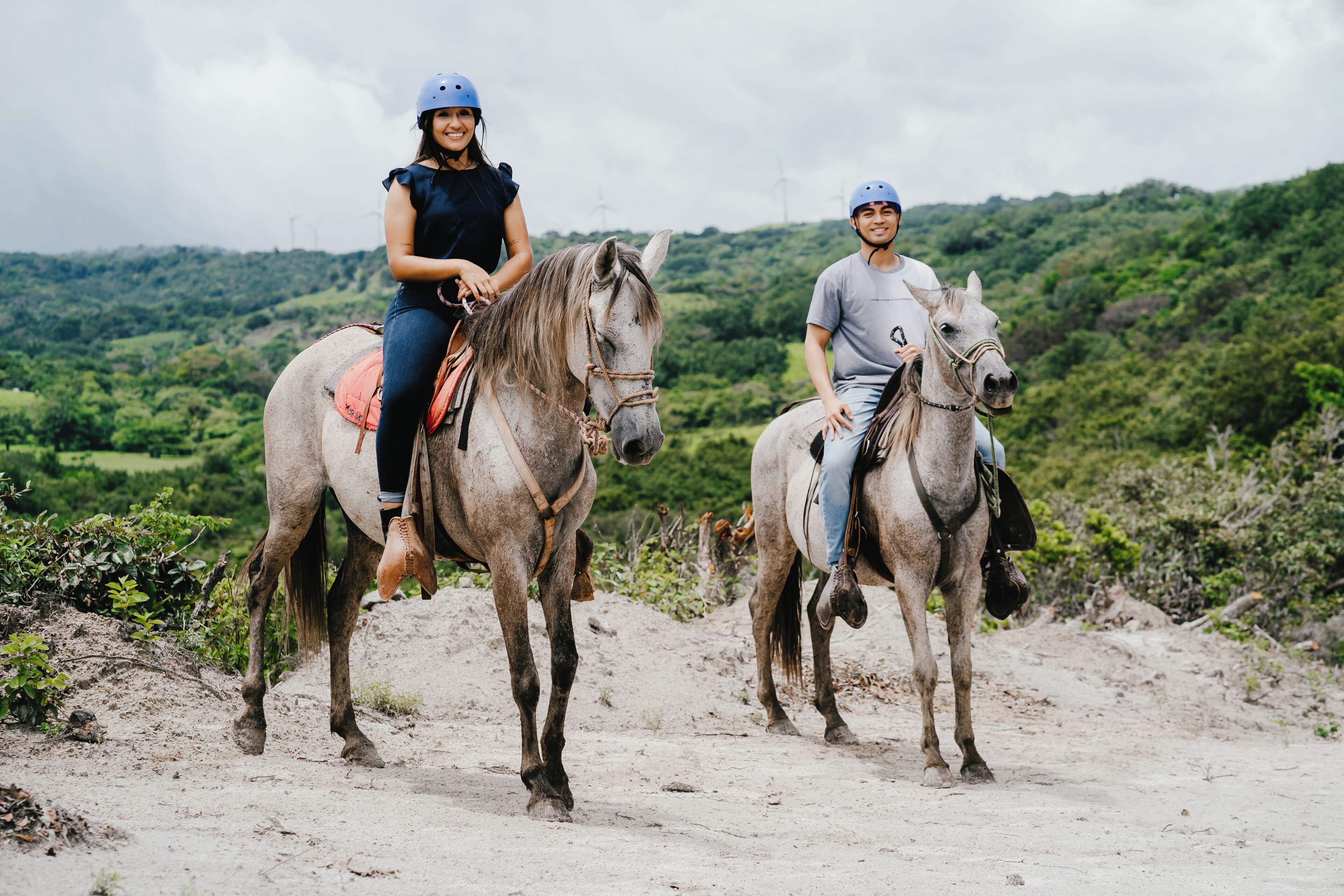 Dues persones cavalquen cavalls per un sender de terra, amb un paisatge verd al fons.