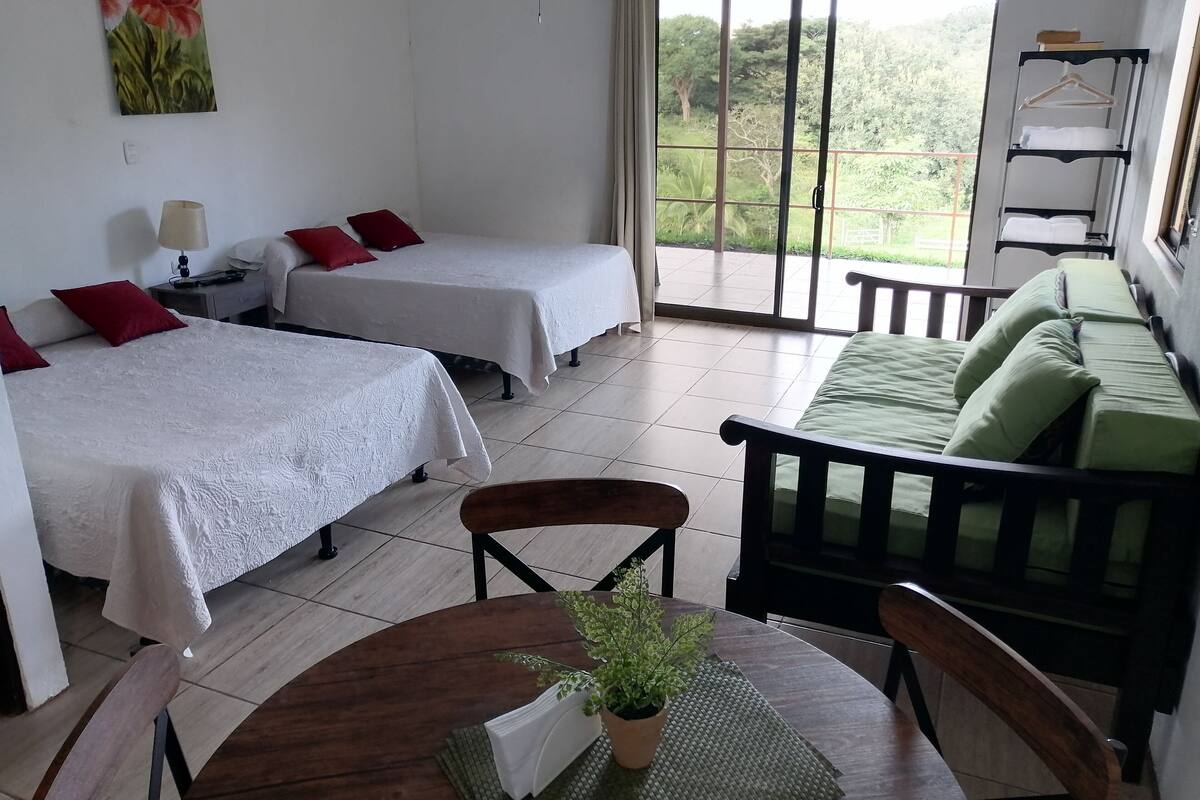A hotel room with two beds, a sofa, a dining table with chairs, a potted plant, and a sliding glass door leading to a balcony.