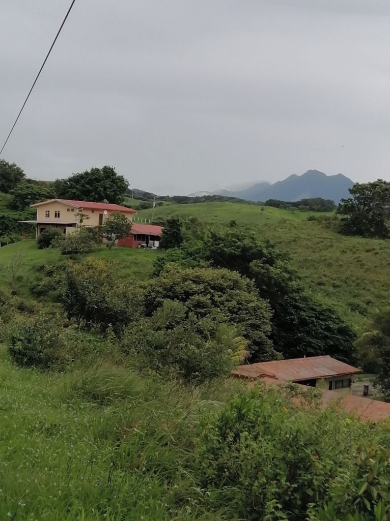 Un paysage rural avec une maison située sur une colline, entourée de verdure et avec d’autres collines en arrière-plan.