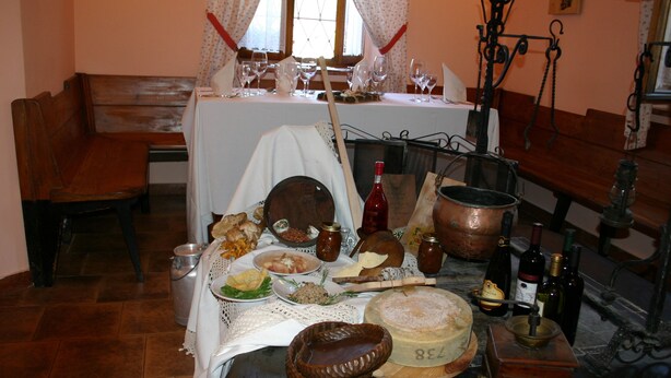 A dining table set with various food items, bottles, and a large cheese wheel. - Albergo Alle Alpi (Comeglians)