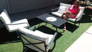 Un patio con dos sillas blancas acolchadas, una mesita y una zona de césped artificial verde.