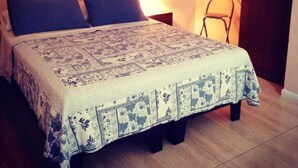 Una cama con una colcha estampada, dos almohadas, una mesita de noche con una lámpara y una puerta a la derecha.