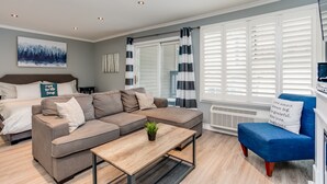 Una sala de estar moderna con un sofá modular, un sillón azul, una mesa de centro de madera y un ventanal con contraventanas blancas.