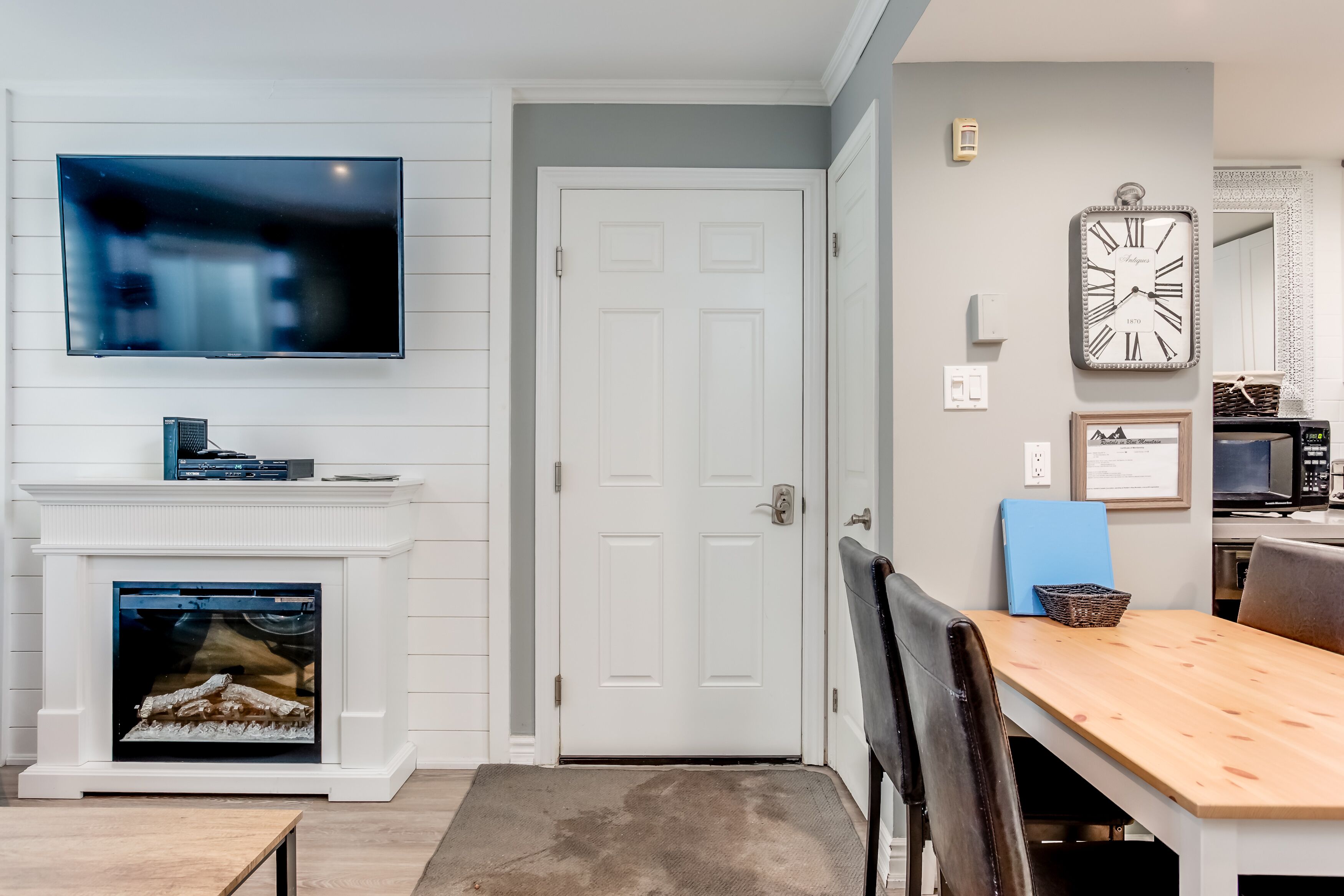 A modern living room with a flat-screen TV mounted above a fireplace, a dining area with a wooden table and chairs, and a wall-mounted clock.