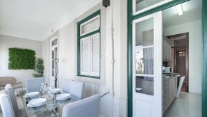Comedor con mesa de vidrio, platos blancos y sillas grises. Se observa una decoración verde en la pared y una vista hacia la cocina.