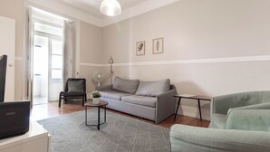 Una sala de estar con un sofá gris, un sillón verde, una mesita con una lámpara y un ordenador portátil sobre un escritorio.