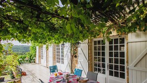 Une terrasse extérieure avec une table dressée pour le repas, entourée de chaises et surmontée d’une pergola couverte de plantes grimpantes.
