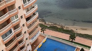 A high-rise building with multiple balconies overlooking a blue swimming pool and a beach.
