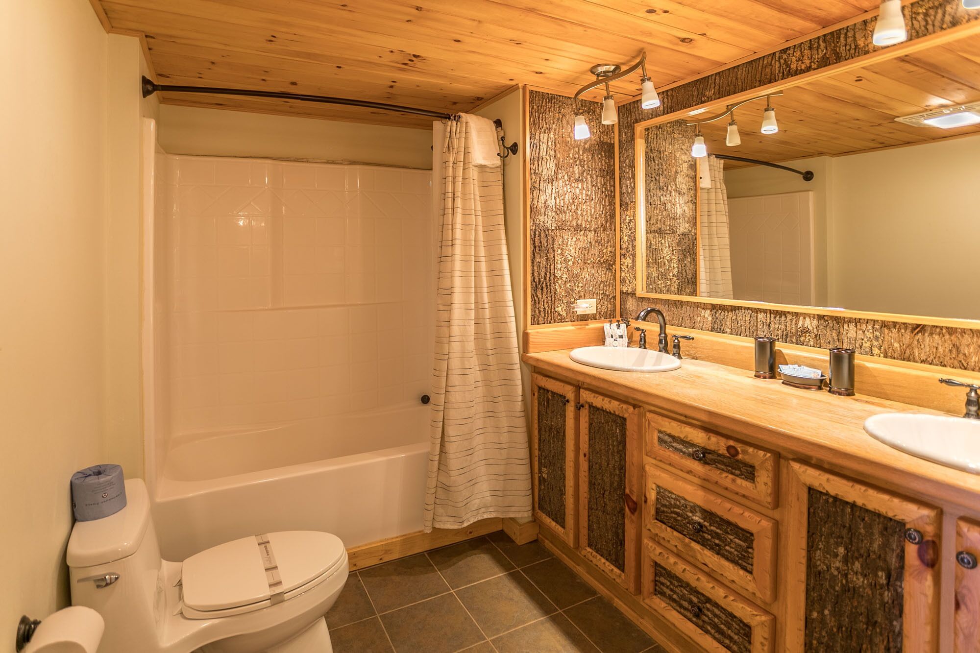Un baño con techo de madera, una bañera, un cortinón de ducha, un inodoro y un tocador con espejo y dos lavamanos.