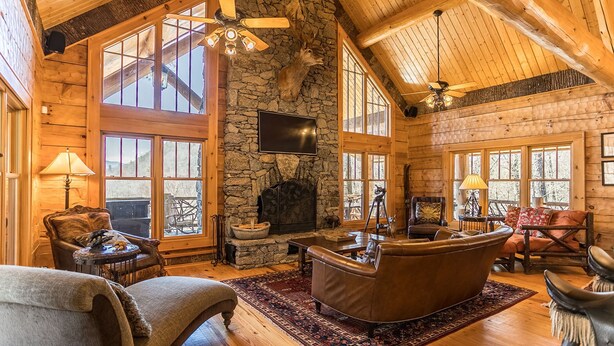 A log cabin interior with a stone fireplace, large windows, and wooden ceiling beams. - Red Stag Lodge at Eagles Nest: Hot tub, game room, outdoor fireplace, summer concerts (Banner Elk)