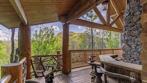 A wooden deck with a stone wall, wooden chairs, and a table. - Red Stag Lodge at Eagles Nest: Hot tub, game room, outdoor fireplace, summer concerts (Banner Elk)