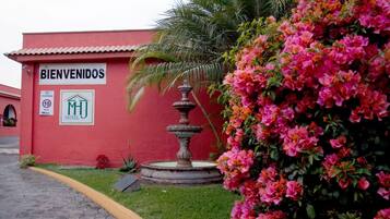 A red building with a 'BIENVENIDOS' sign, a fountain, and pink flowers.