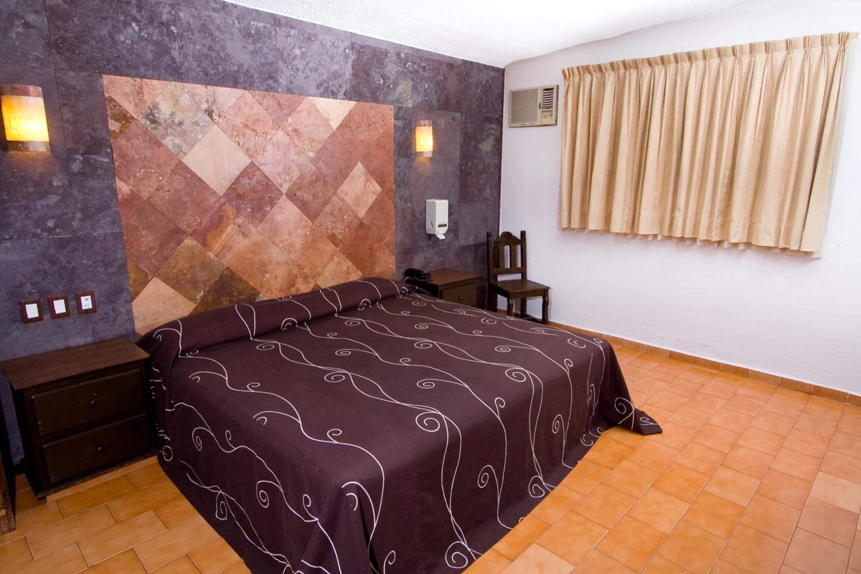 Un dormitorio con cama, mesita de noche, silla, aire acondicionado instalado en la pared y ventana con cortinas.