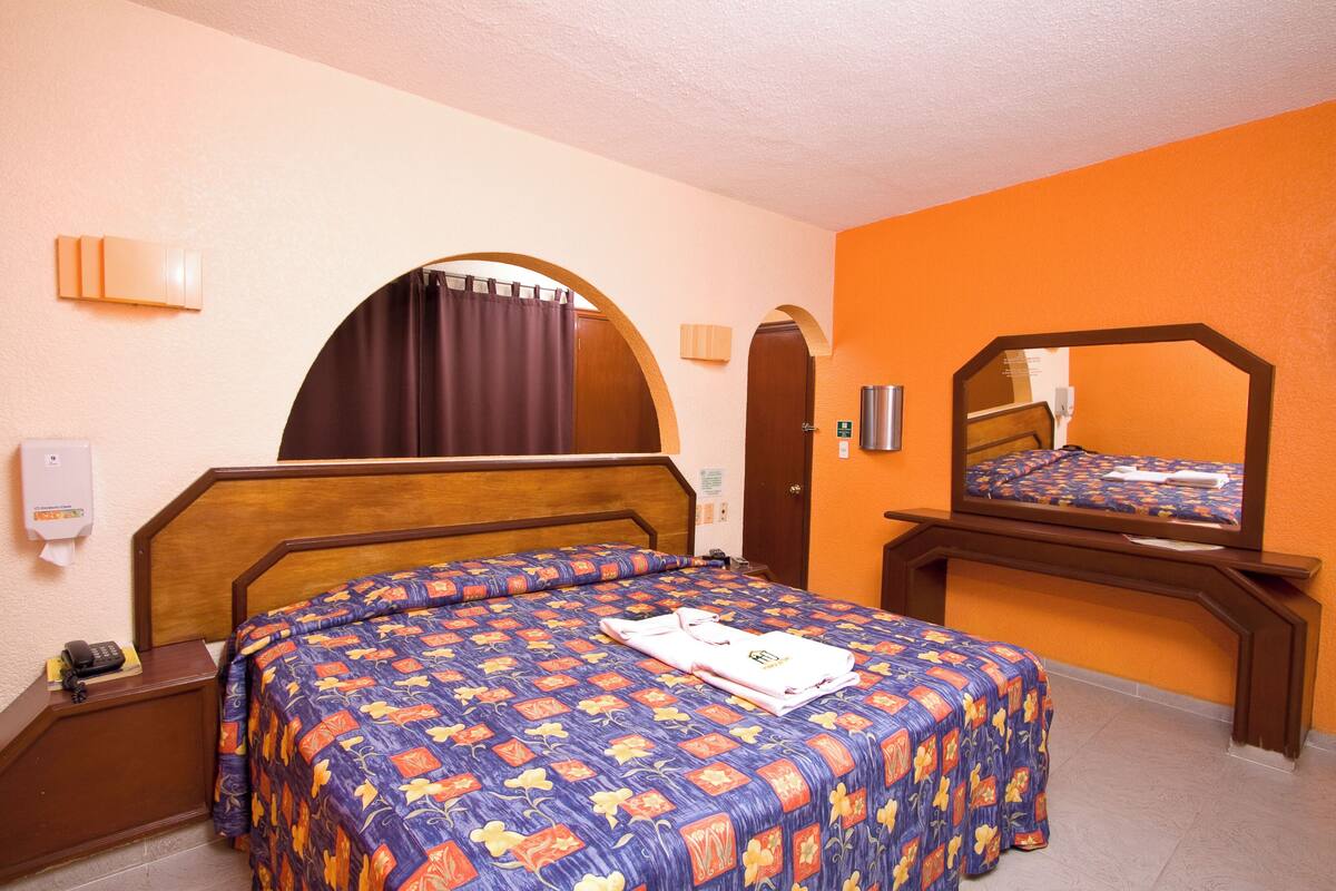 Ett hotellrum med en säng i trä, orange väggar, en spegel och en telefon på nattduksbordet.