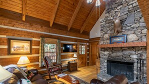 Un acogedor interior de cabaña de madera con chimenea de piedra, muebles de cuero y un televisor en la pared.