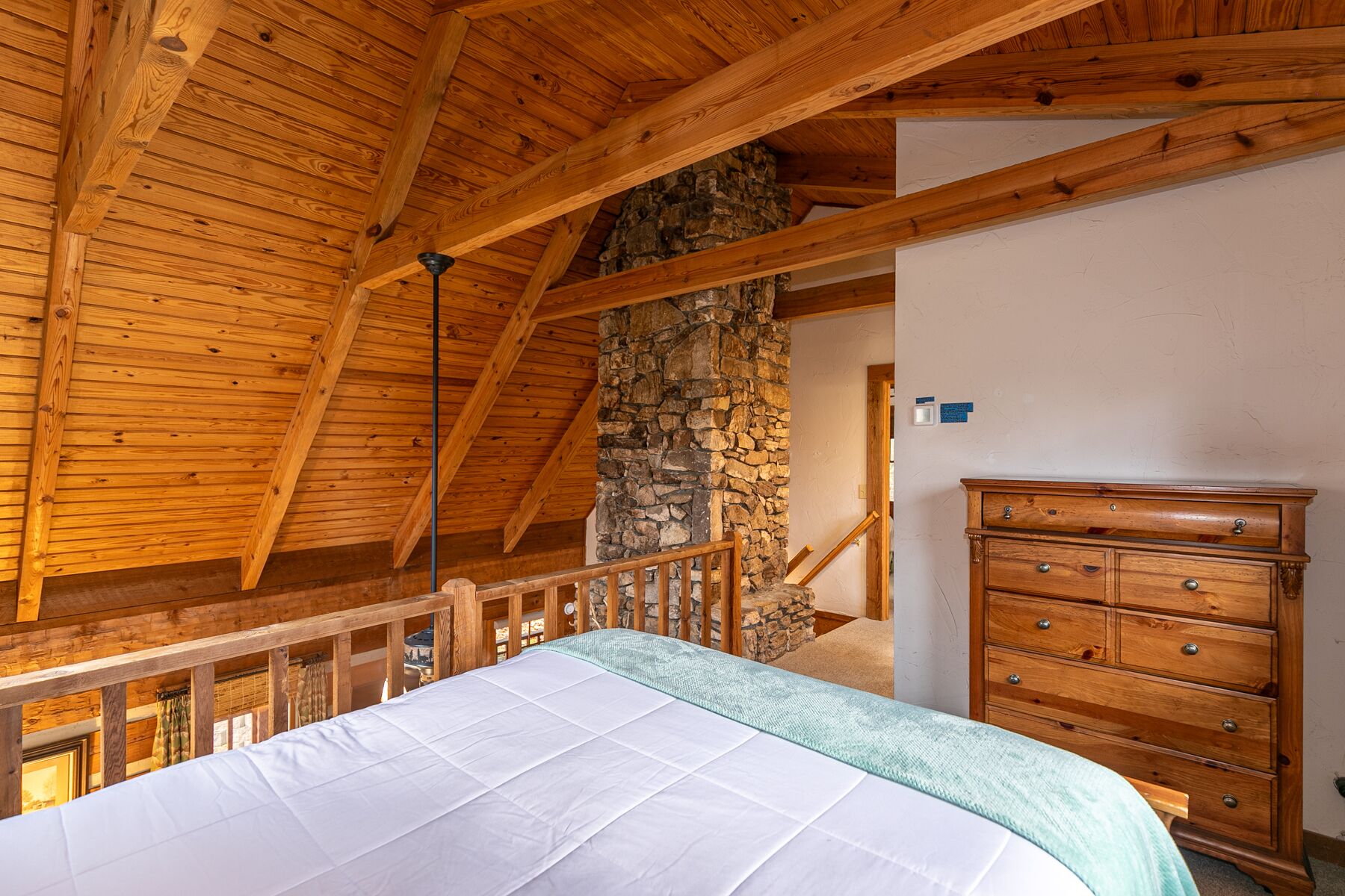 A bedroom with a wooden ceiling, a stone wall, a bed with a white mattress, and a wooden dresser.
