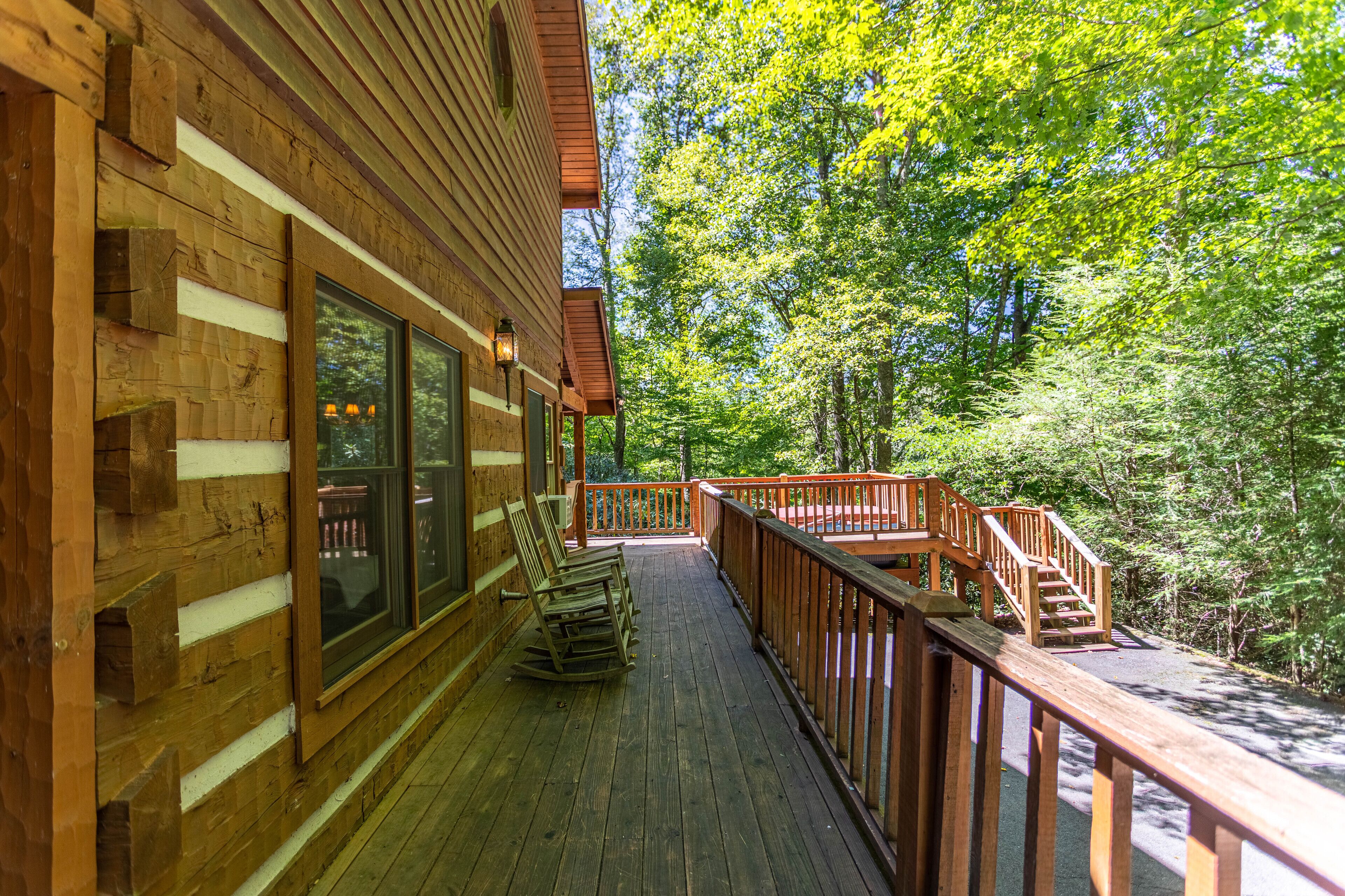 A wooden cabin with a deck, rocking chairs, and a staircase leading to a deck area surrounded by trees.