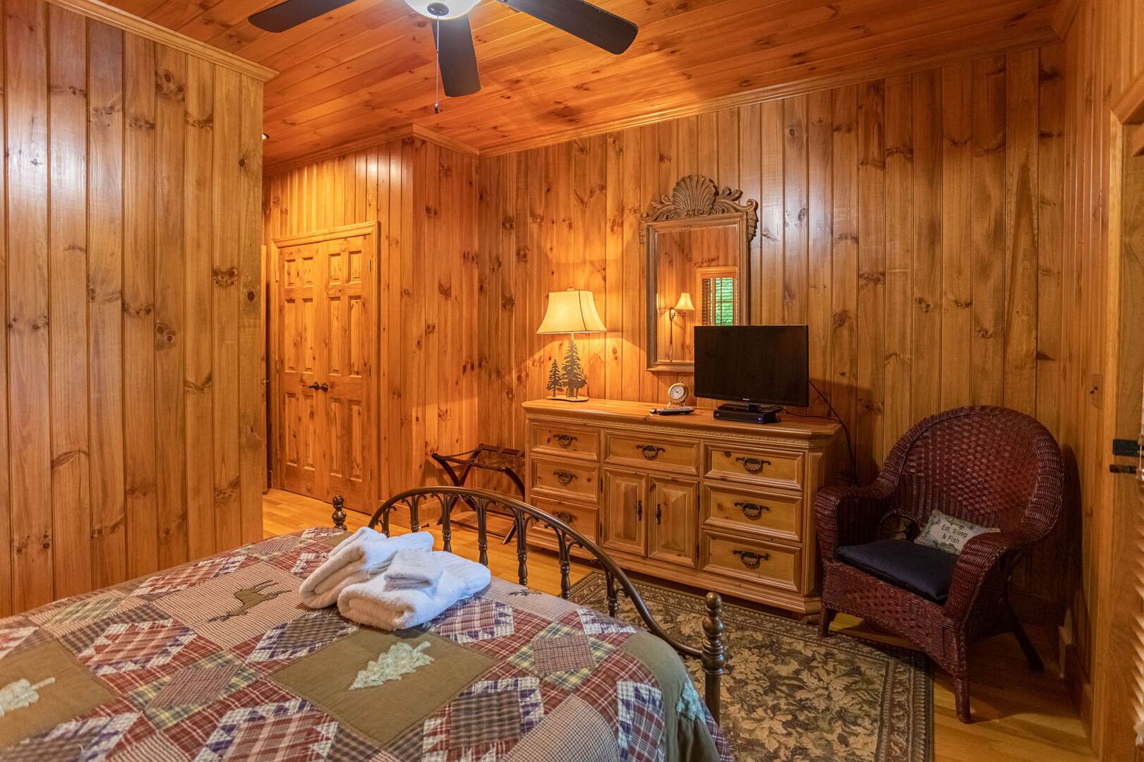 A wooden-paneled bedroom with a bed, dresser, chair, and ceiling fan.